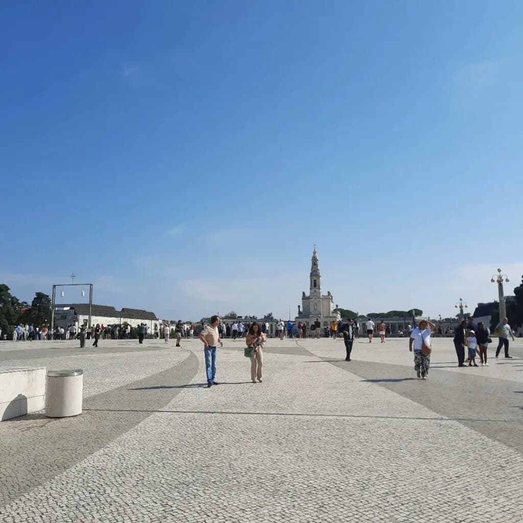 Vista panorâmica da Esplanada do Santuário de Fátima sob um céu ensolarado com fiéis a caminhar em reflexão.